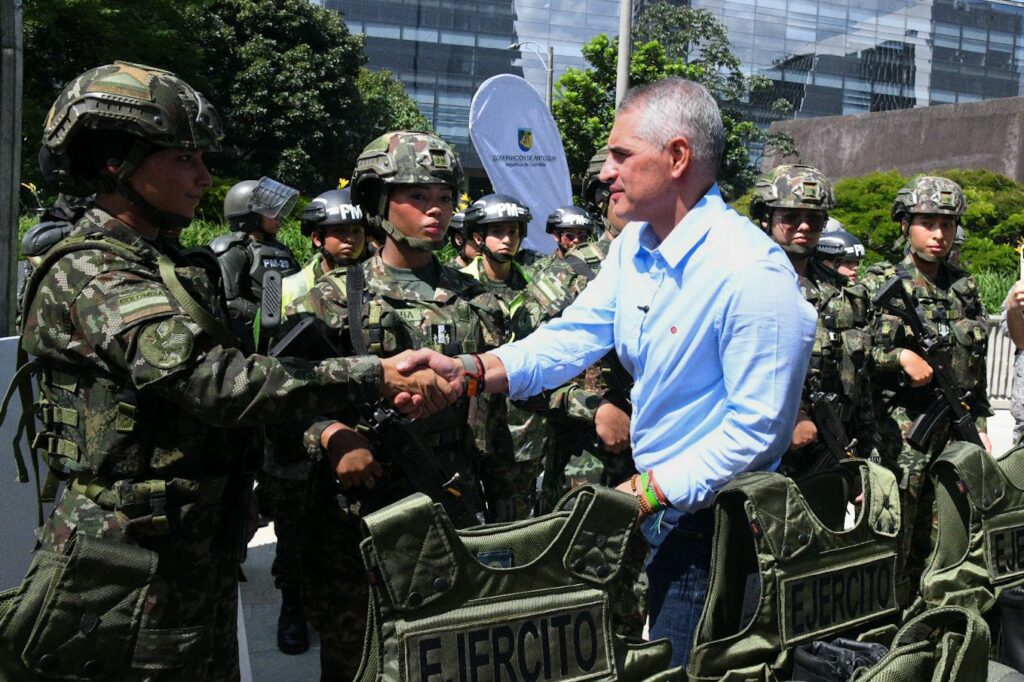 "El Gobernador de Antioquia, Andrés Julián, estrecha la mano de una mujer soldado del Ejército Nacional durante un evento oficial de entrega de dotación. En primer plano se ven chalecos antibalas con el rótulo 'EJÉRCITO' y otros militares uniformados al fondo."