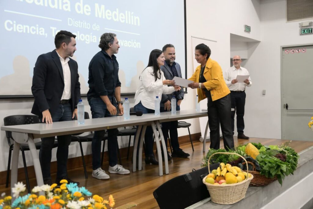 Representantes de la Alcaldía de Medellín entregan un certificado a una mujer frente a una canasta de productos agrícolas frescos.