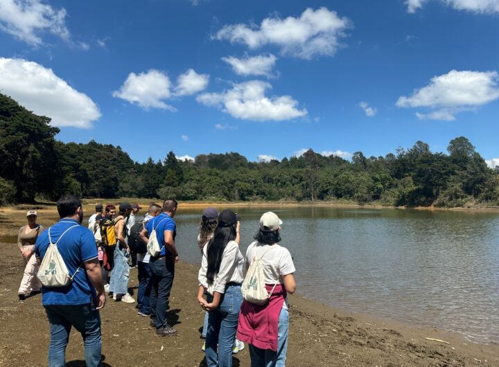 Grupo de turistas a la orilla de una represa rodeada de bosque denso bajo un cielo azul con nubes en el Parque Arví, Medellín.