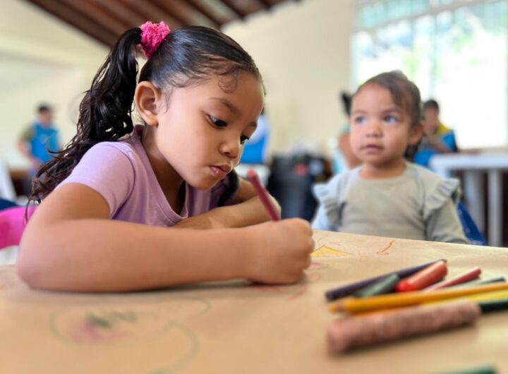 Una niña pequeña concentrada dibujando con colores en un aula de Buen Comienzo, con otra niña al fondo, representando la atención integral en Medellín.