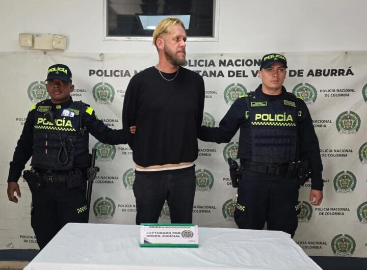Un ciudadano extranjero bajo custodia, escoltado por dos agentes de la Policía Nacional de Colombia frente a un muro institucional.