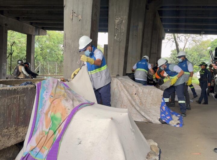 Personal de limpieza con chalecos azules, cascos blancos y guantes trabajando en el desmonte de cambuches y recolección de residuos bajo un puente vehicular en Medellín, acompañados por la Policía Nacional.