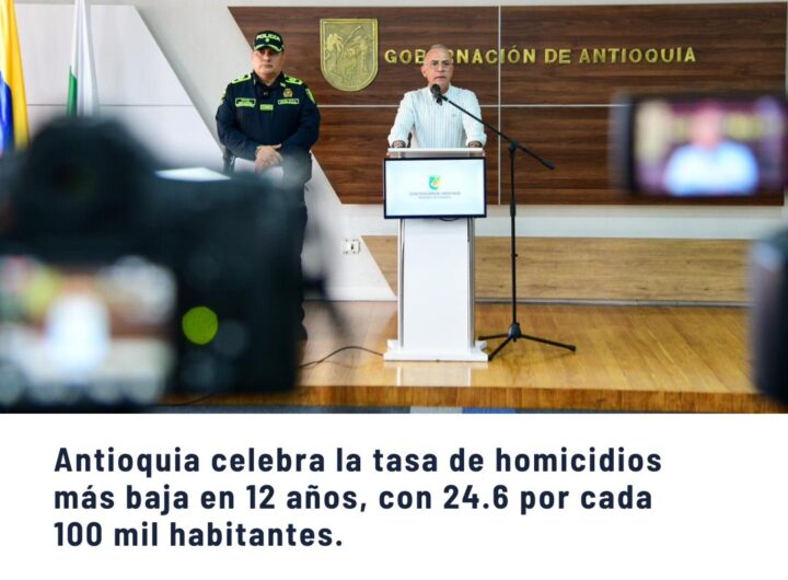 Conferencia de prensa con orador en podio y policía, anuncio de Antioquia celebrando la tasa de homicidios más baja en 12 años con 24.6 por cada 100 mil habitantes.
