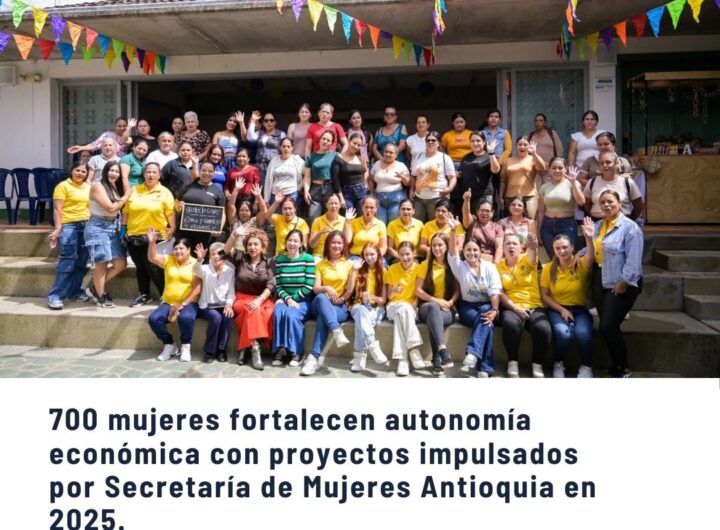 Grupo grande de mujeres sonrientes posando en escaleras al aire libre con banderas de colores, celebrando el fortalecimiento de la autonomía económica de 700 mujeres en Antioquia durante 2025 por la Secretaría de Mujeres.