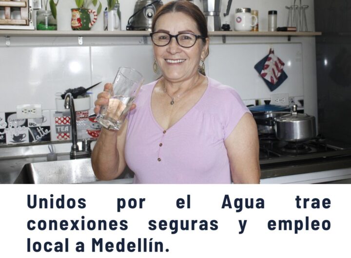 Mujer sonriente sosteniendo un vaso de agua en una cocina, promoviendo el programa Unidos por el Agua que trae conexiones seguras y empleo local a las comunas de Medellín.
